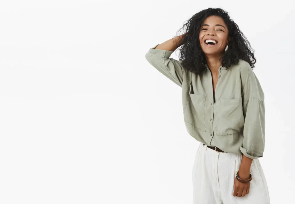Jeune femme souriante, habillée de manière décontractée avec une chemise verte et un pantalon blanc, posant avec naturel sur fond blanc.