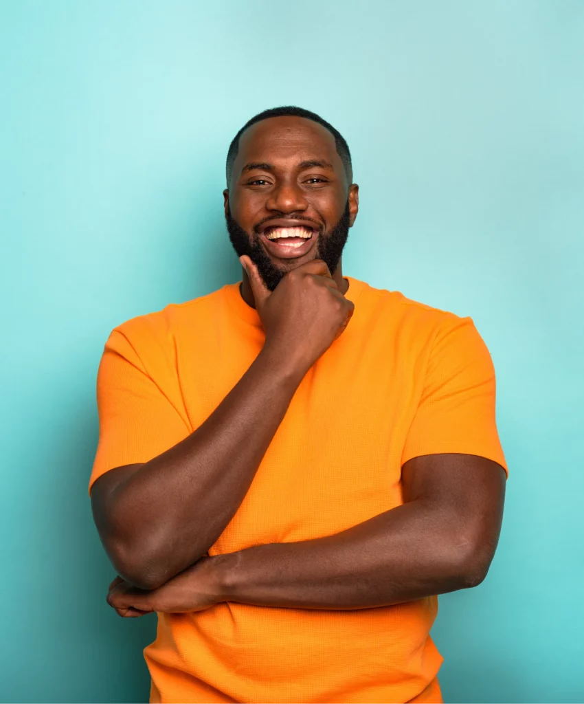 Homme graphiste souriant, vêtu d’un t-shirt orange, posant avec confiance et humour devant un fond bleu vif.