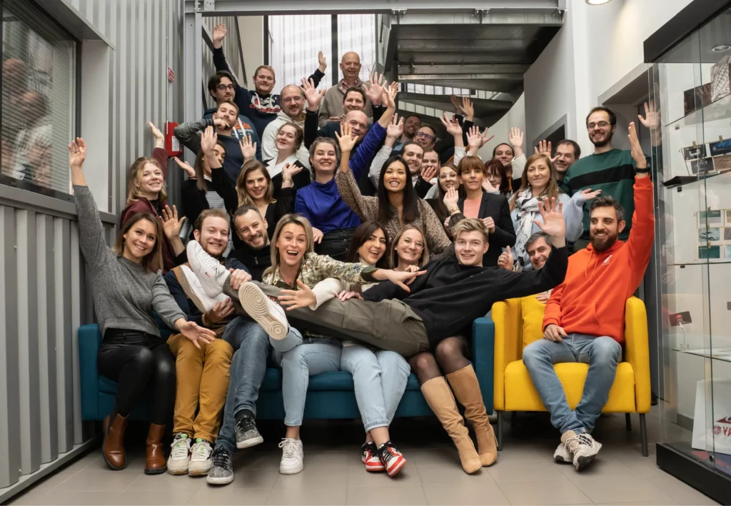 Groupe de collaborateurs de l'agence de 3ma group réunis dans un cadre professionnel, souriants et levant les bras avec énergie, assis sur des canapés dans un espace d’accueil convivial.